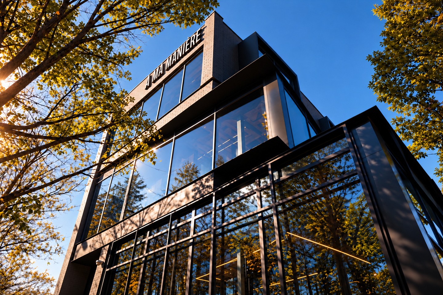Dark brick and glass facade of A Ma Maniere Chicago with the name in black letters, framed by golden autumn trees against a blue sky