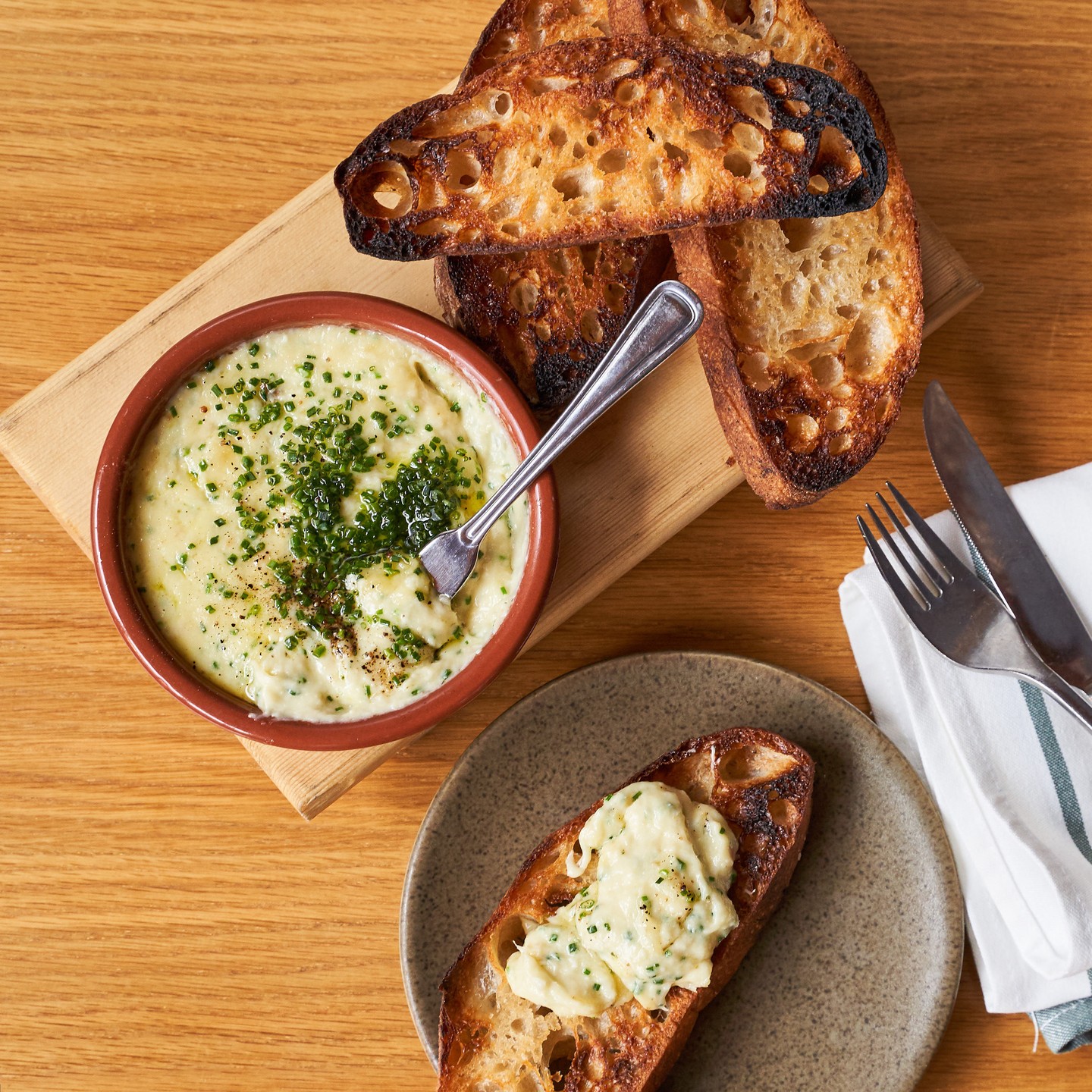 A bowl of potato and salted cod brandade topped with chives beside grilled garlic crostini on a wood board