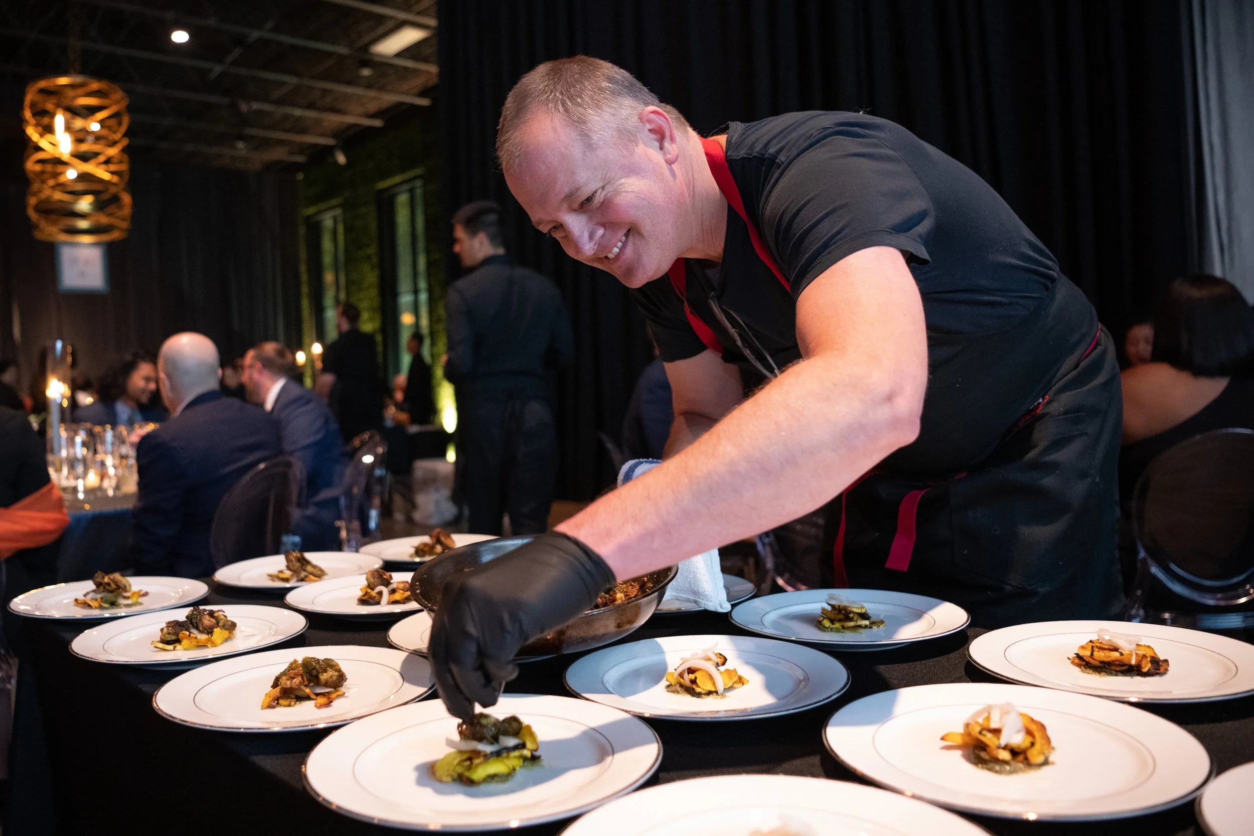 A smiling chef in black gloves plating a course on a row of white plates beside the tables at Celebrity Chef Ball Chicago
