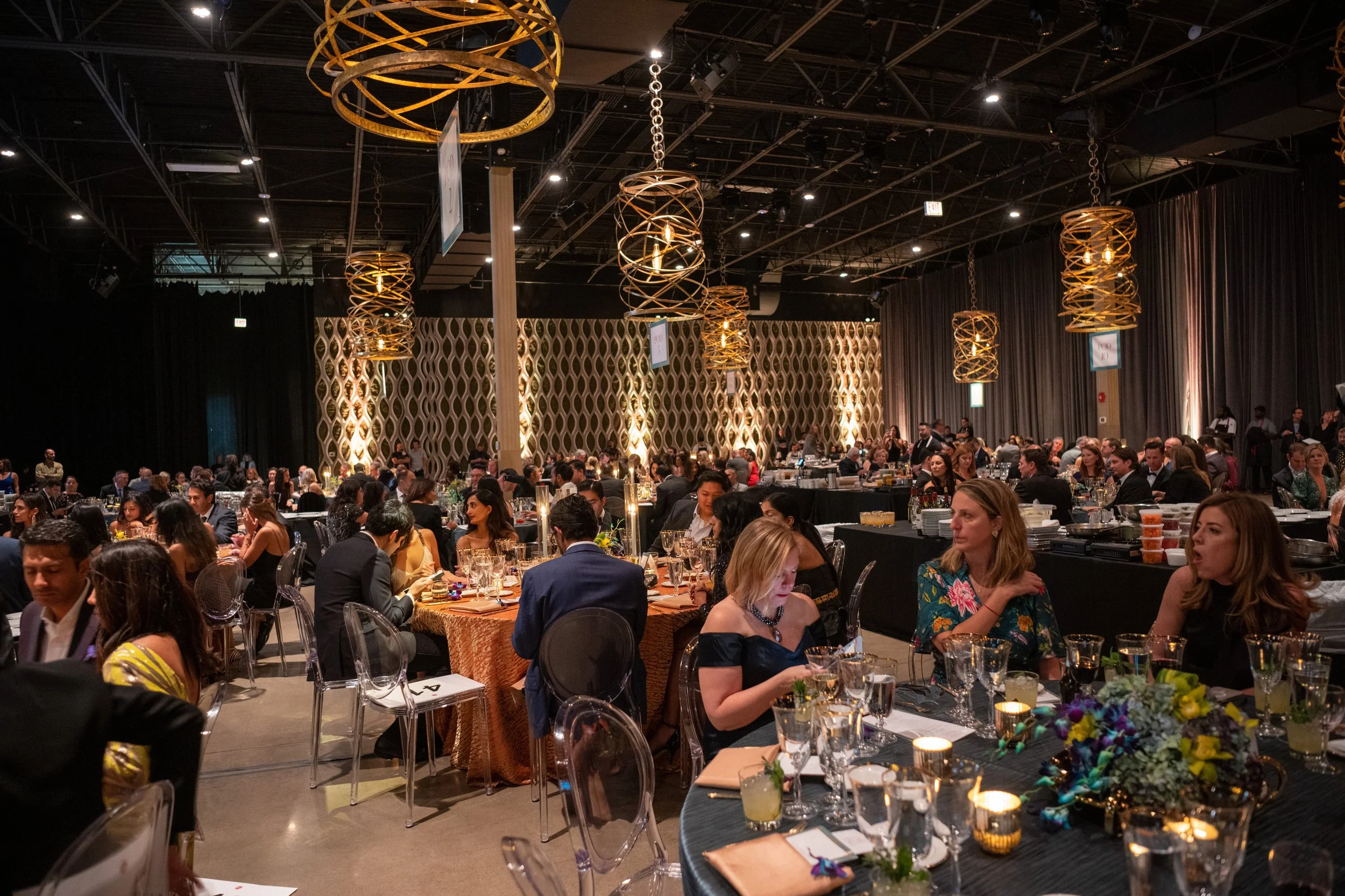 Guests seated at candlelit round tables under gold woven chandeliers in the dining room at Celebrity Chef Ball Chicago