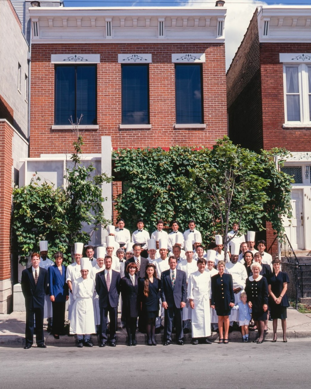 The Charlie Trotter's kitchen and dining room team in whites and suits lined up on the sidewalk in front of the brick building