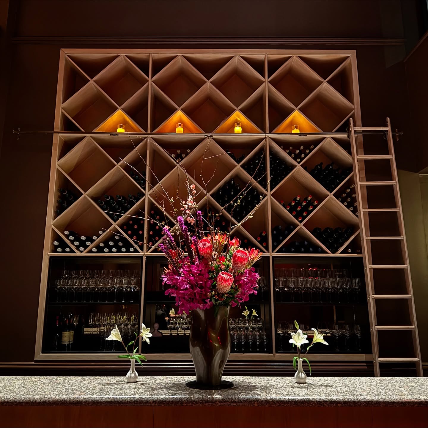 A lit diamond-grid wine wall with a rolling ladder behind a stone counter set with pink flowers