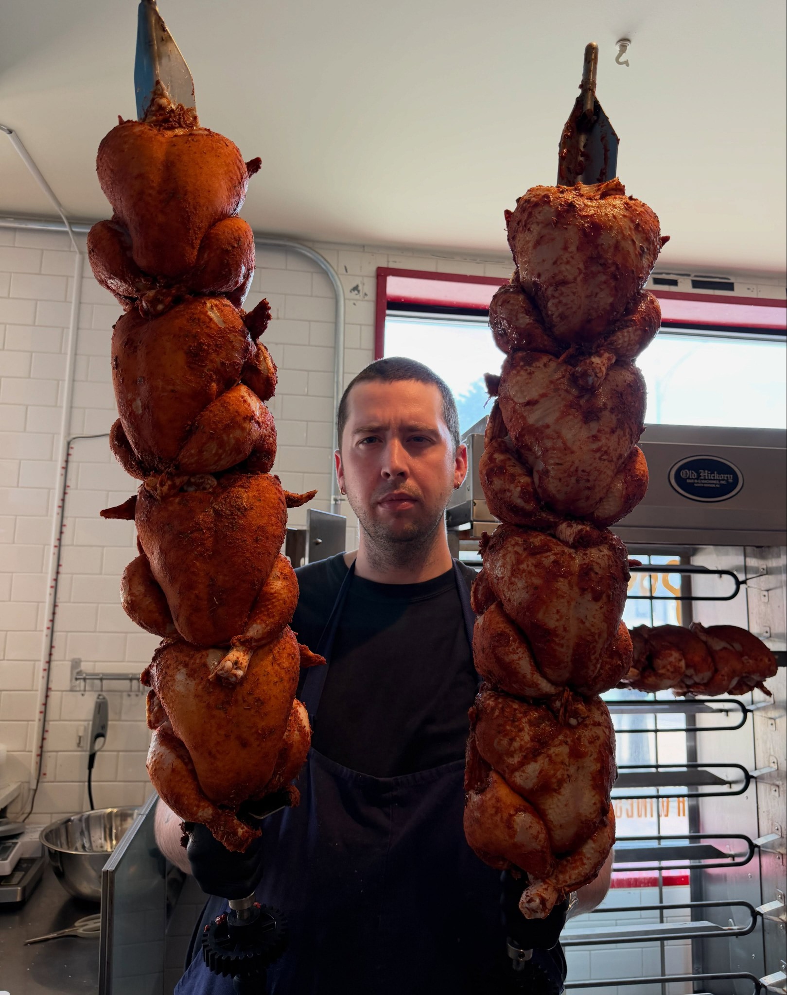 Chef Keagan Beresford holding two spits of seasoned chickens beside the rotisserie at Chez Poulet Chicago