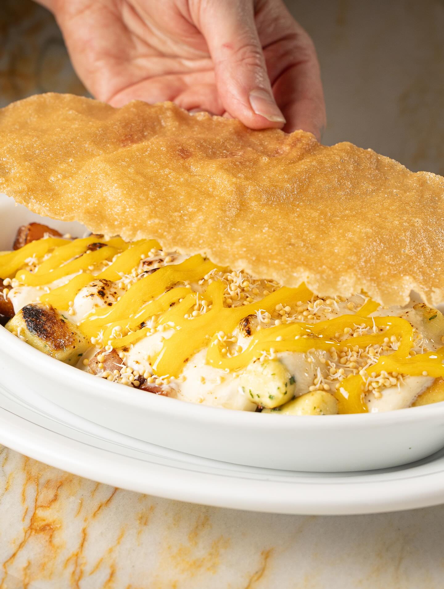 A hand lifts a crisp sheet of brik pastry off Parisian gnocchi with Swiss cheese, ham and egg yolk