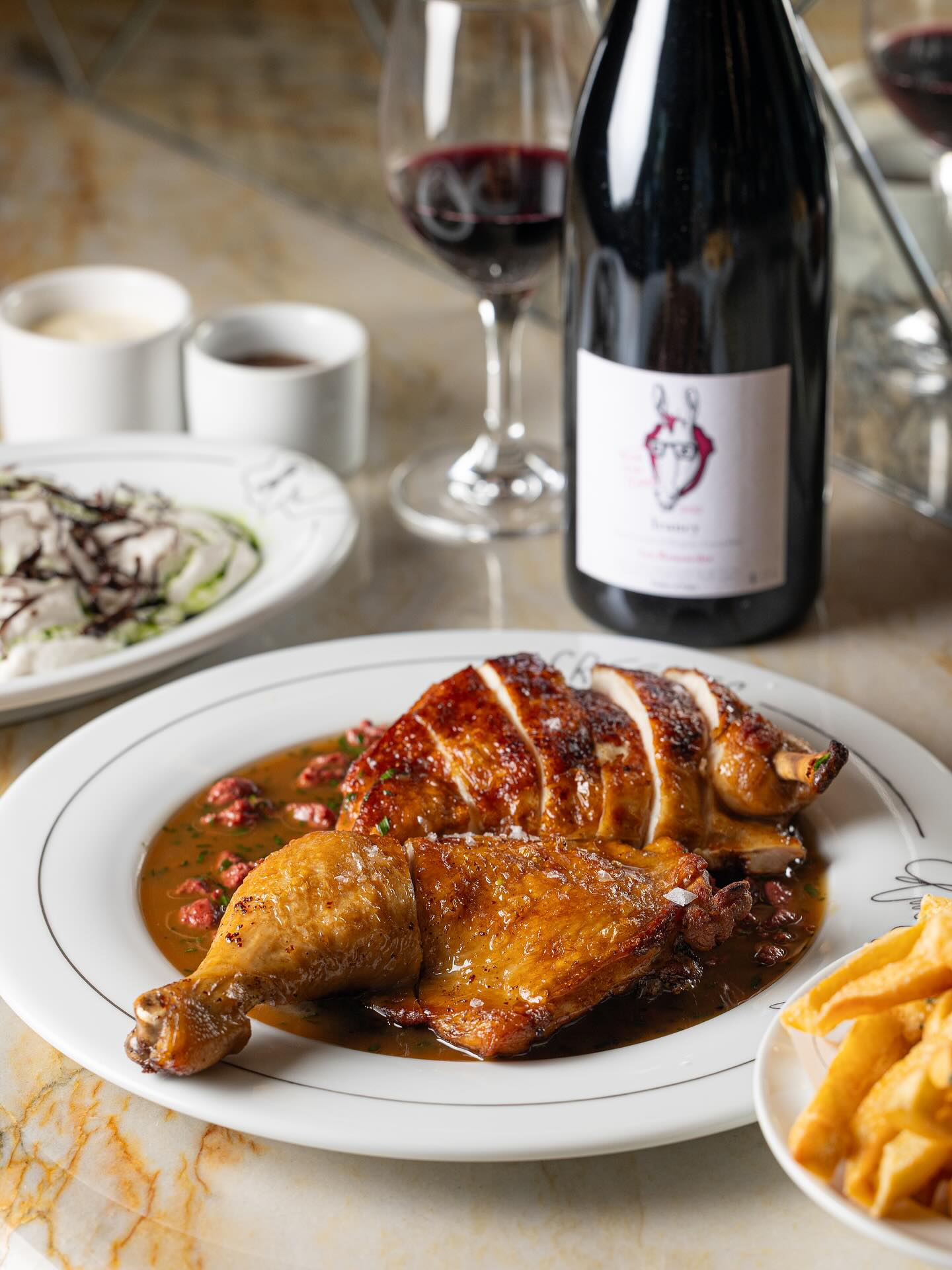 Roasted half chicken in liver and wine sauce with fries and a bottle of red wine on a marble table