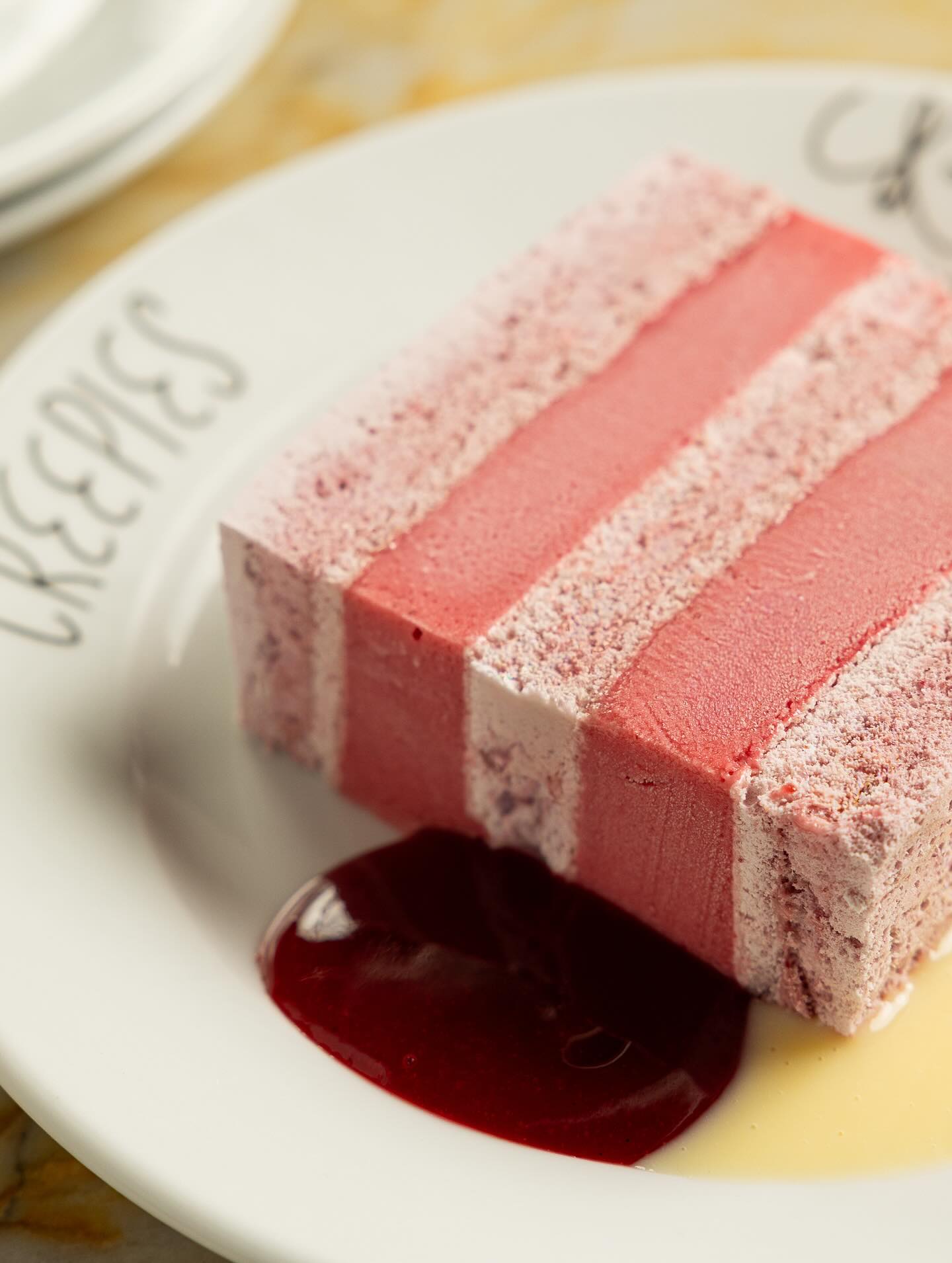 A striped pink slice of sherbet and meringue cake with berry sauce and buttermilk on a white plate