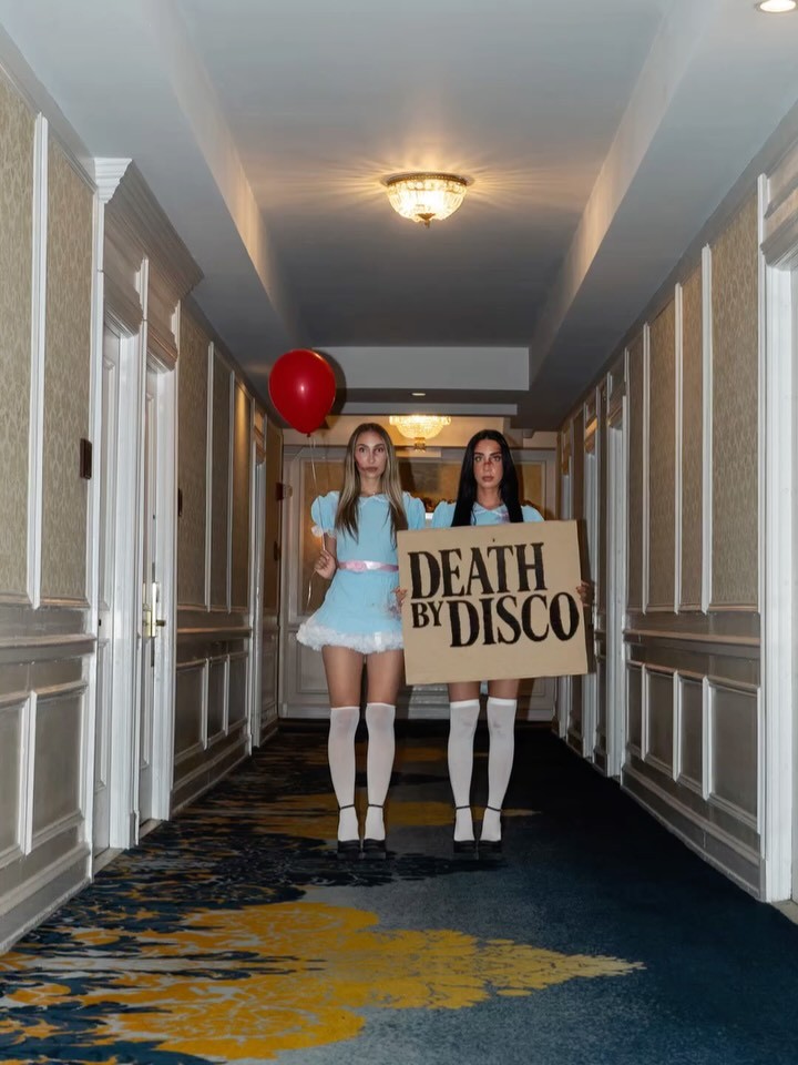 Two performers in matching blue dresses holding a Death By Disco sign in a hotel hallway