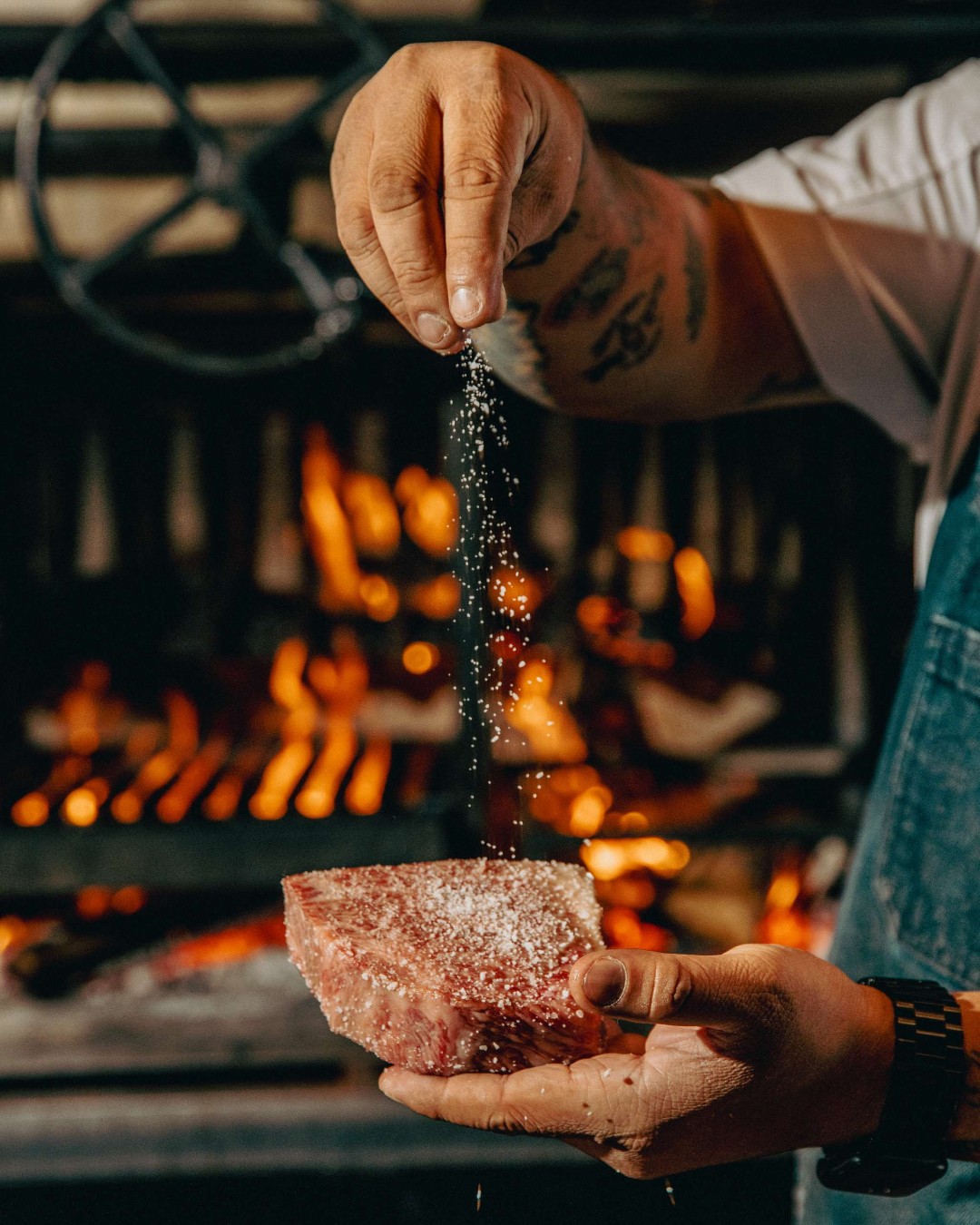 A cook salts a raw steak in front of the glowing wood-fired hearth at El Che