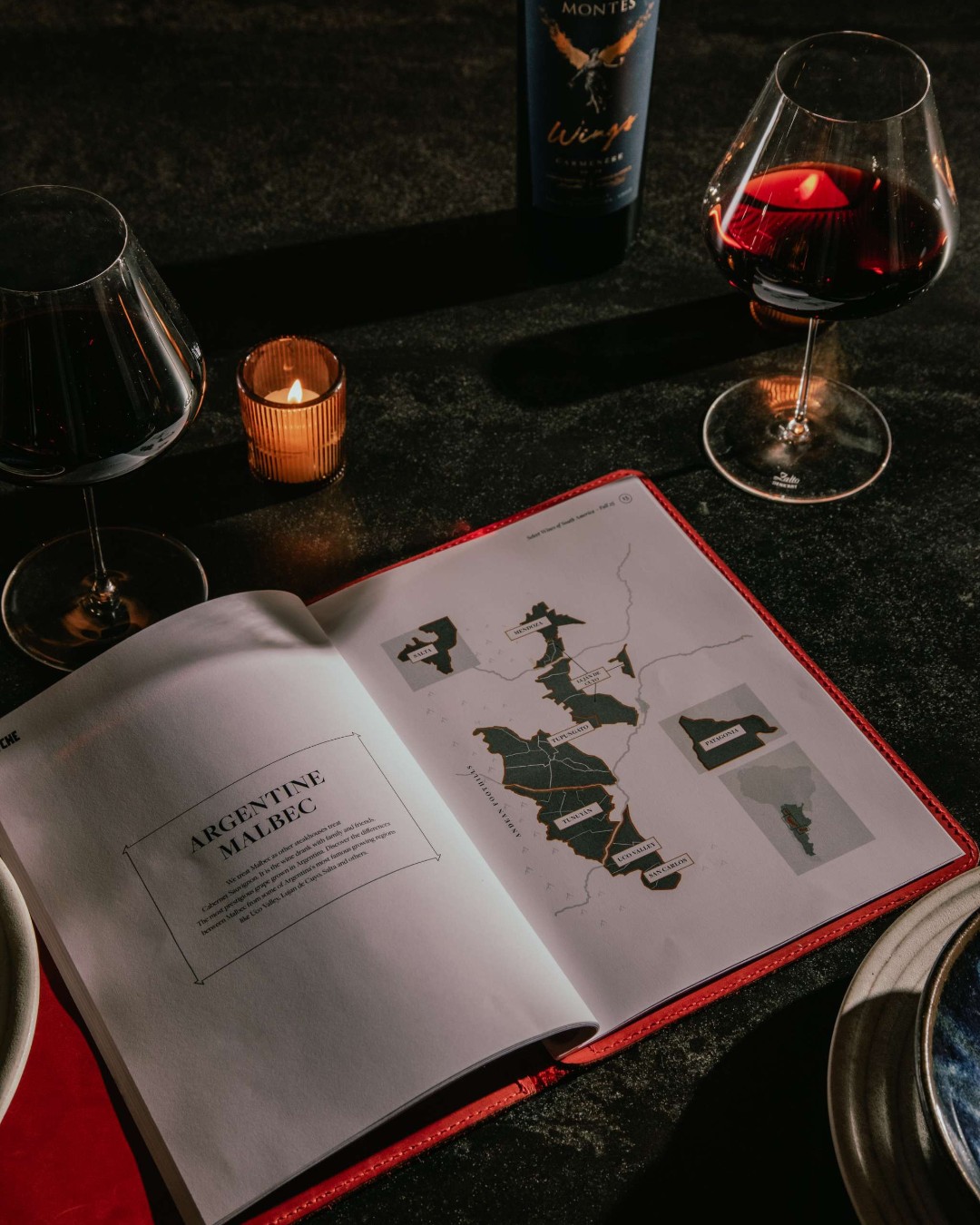 An open wine book with a map of South America beside a glass of red wine and a candle