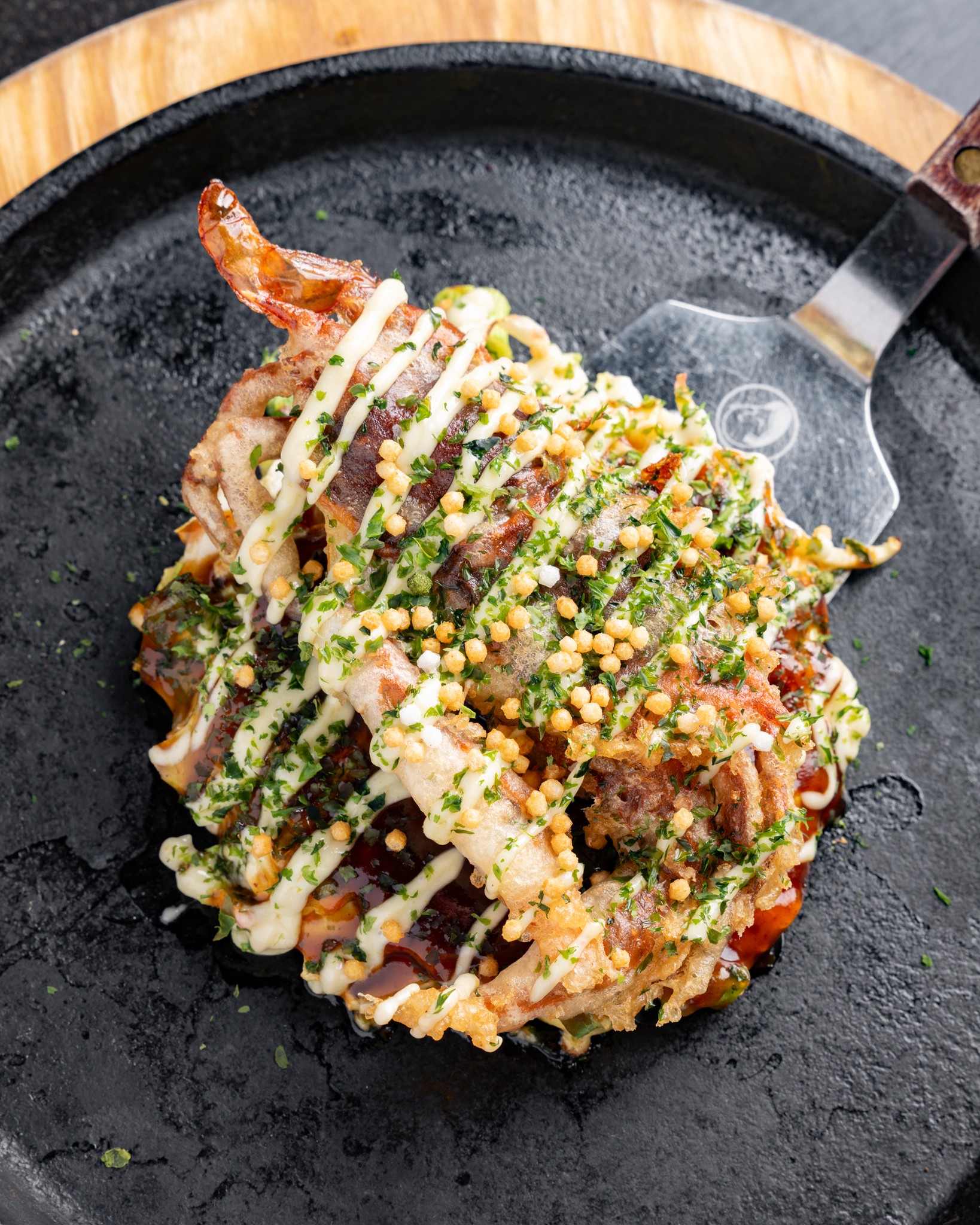 An okonomiyaki topped with mayo, sauce and aonori served on a cast iron teppan with a spatula at Gaijin