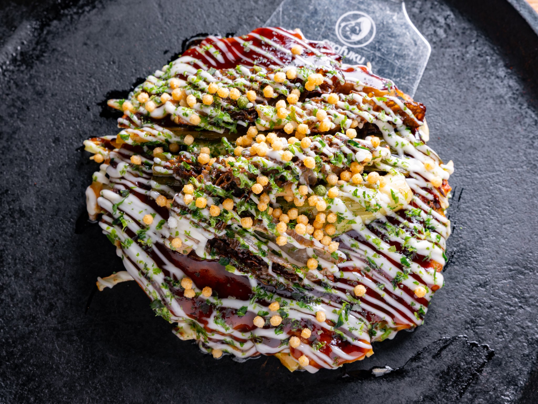 Osaka-style okonomiyaki striped with kewpie mayo and okonomiyaki sauce on a black griddle plate at Gaijin