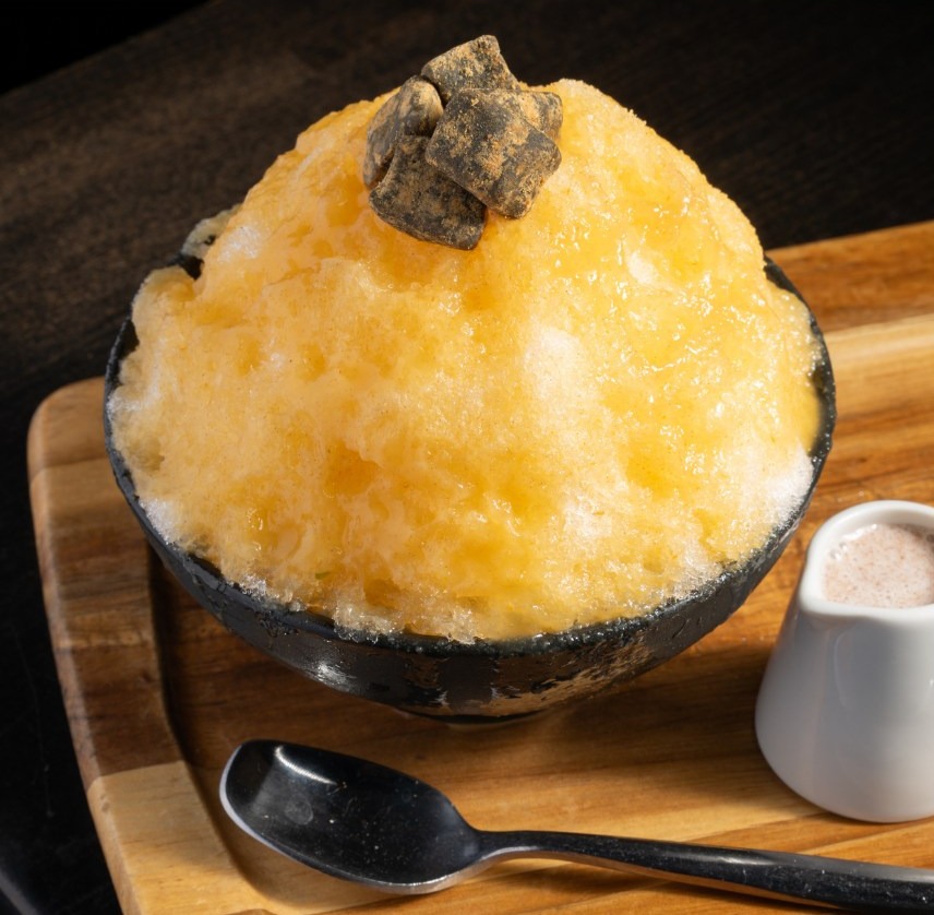 A dome of persimmon shaved ice topped with kinako candy in a dark bowl on a wooden board