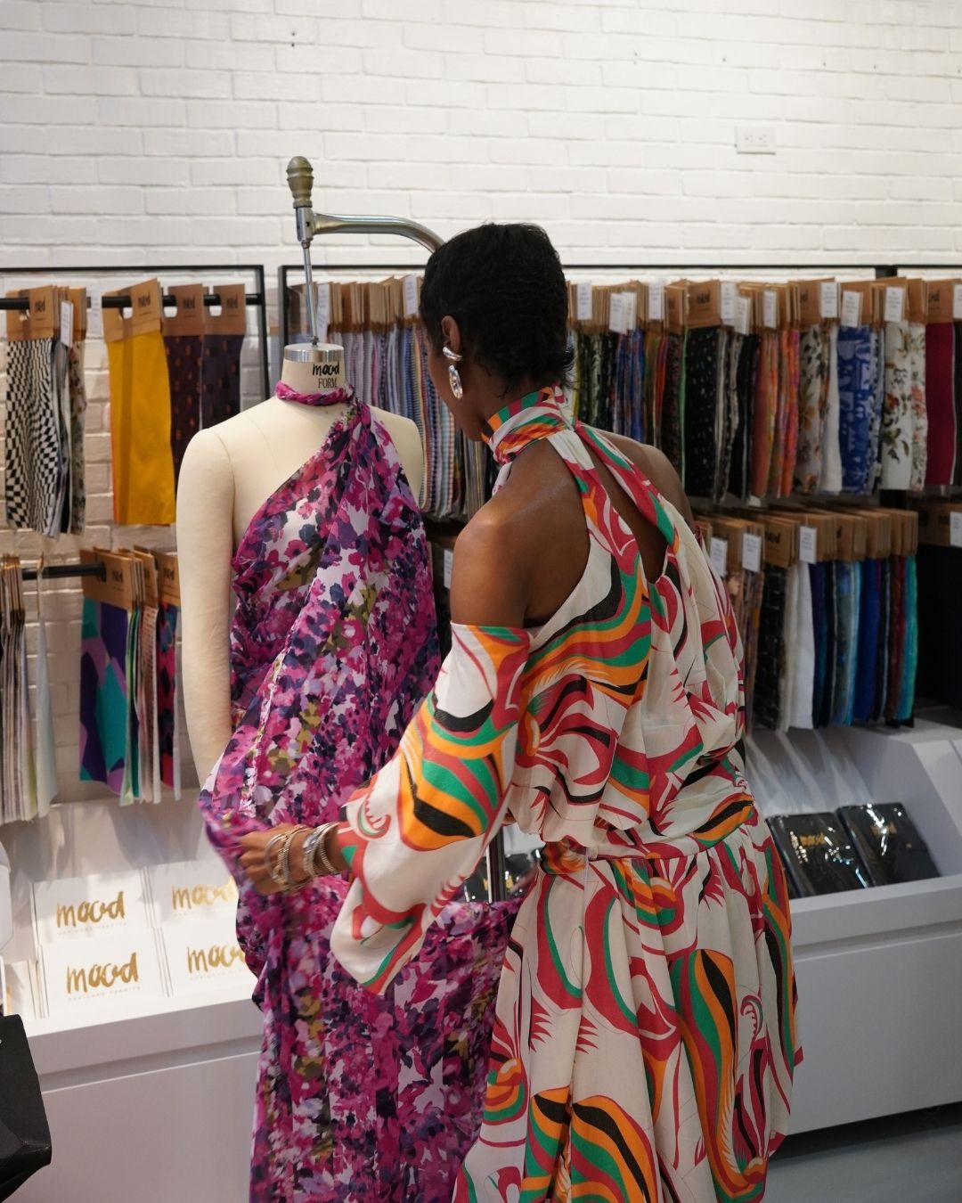 Designer draping patterned fabric on a mannequin in front of shelves of Mood Fabrics bolts