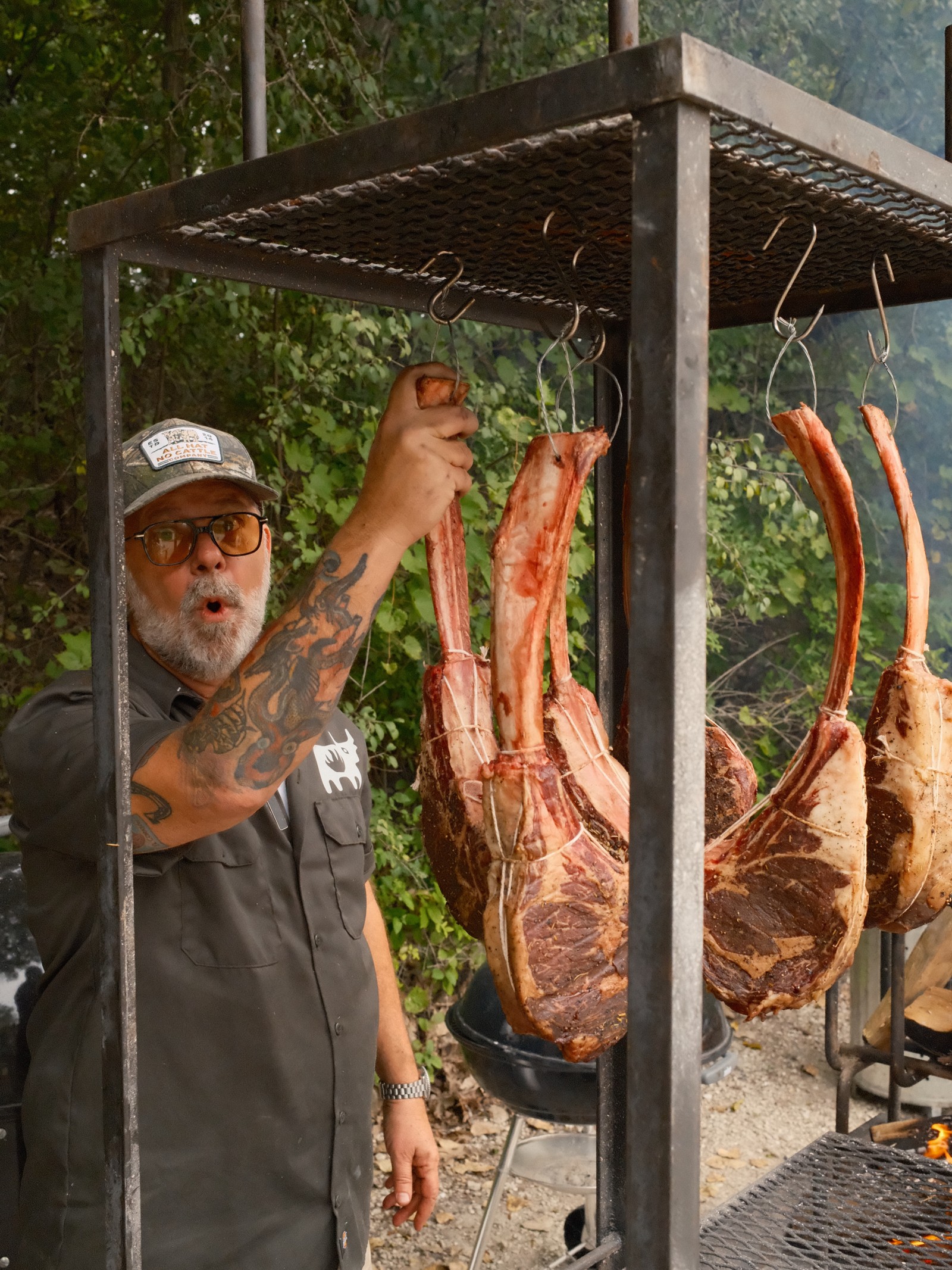 Chef John Manion hangs whole tomahawk steaks over an open fire on a steel rig outdoors