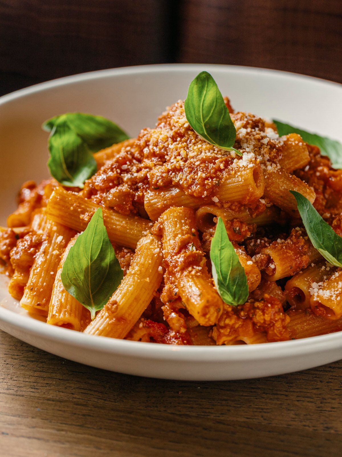 A white bowl of rigatoni in chicken bolognese topped with grated cheese and fresh basil leaves