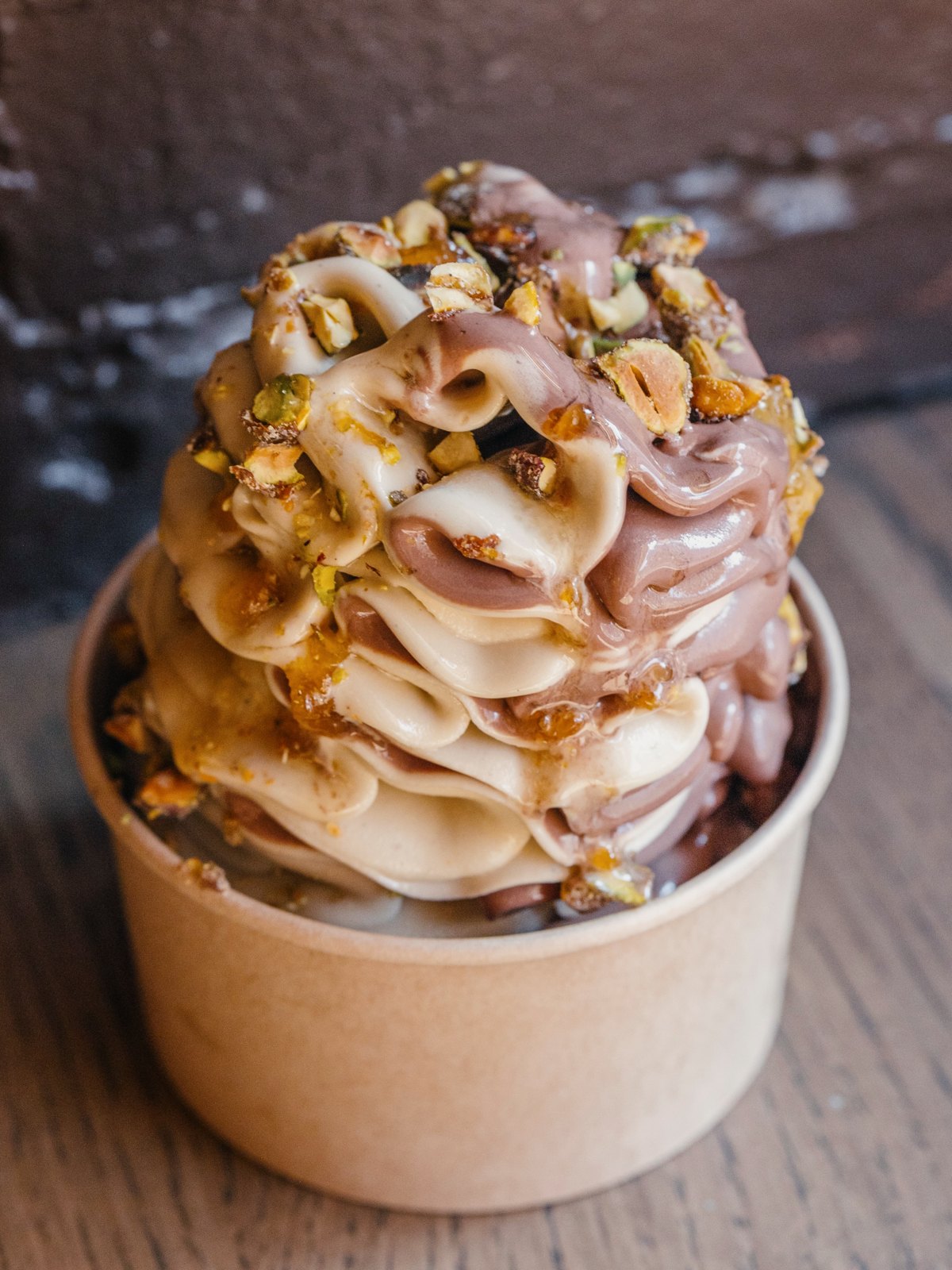 A tall twist of chocolate and pistachio soft-serve gelato topped with pistachio praline in a paper cup