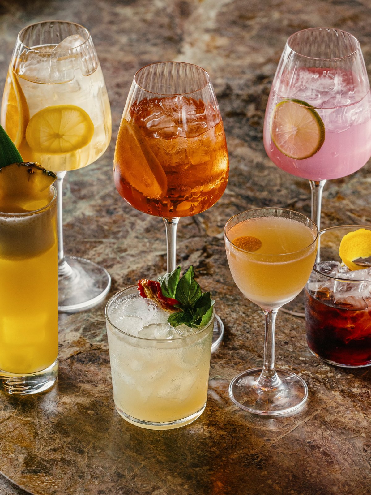 A row of spritzes and cocktails in wine glasses and coupes with citrus garnishes on a stone bar top
