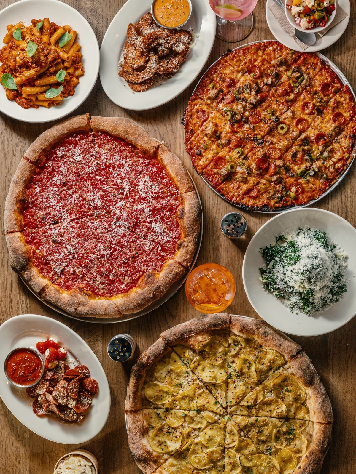 Overhead spread of Chicago Thin and Crispy and New York Crispy pizzas with pepperoni chips, salad, pasta and spritzes on a wood table