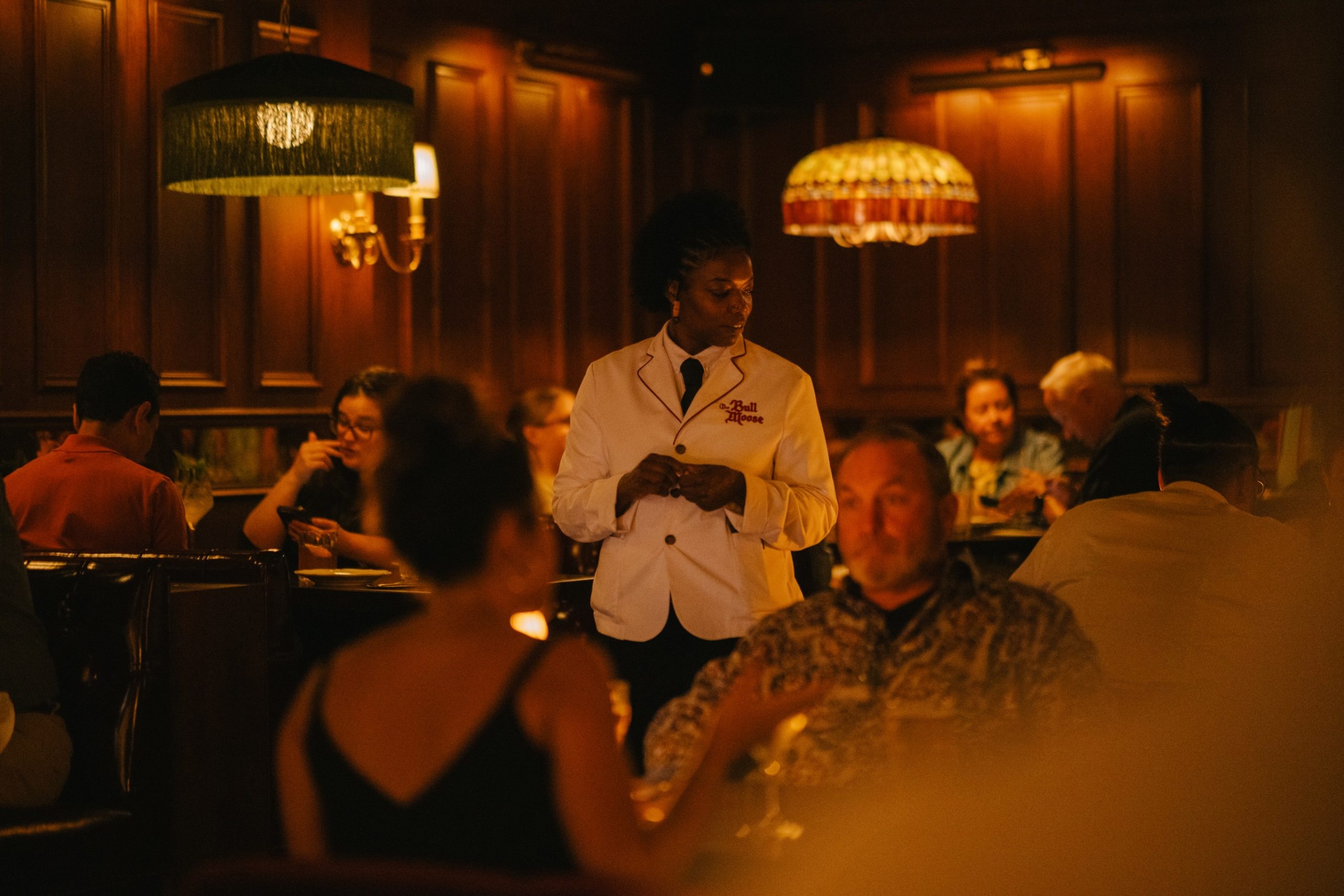 A server in a white jacket attends diners in the dim wood-paneled dining room at The Bull Moose