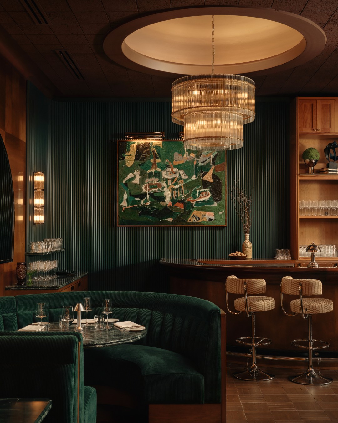 The dining room at The Ives with deep green banquettes, a crystal chandelier, a painting and the bar in the background
