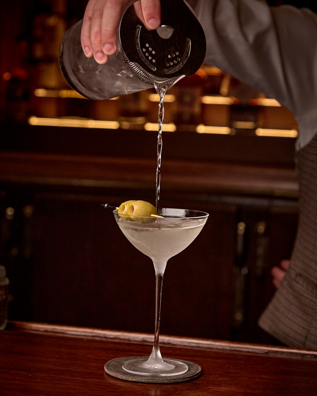 A bartender strains a martini from a shaker into a stemmed glass with an olive at The Ives bar