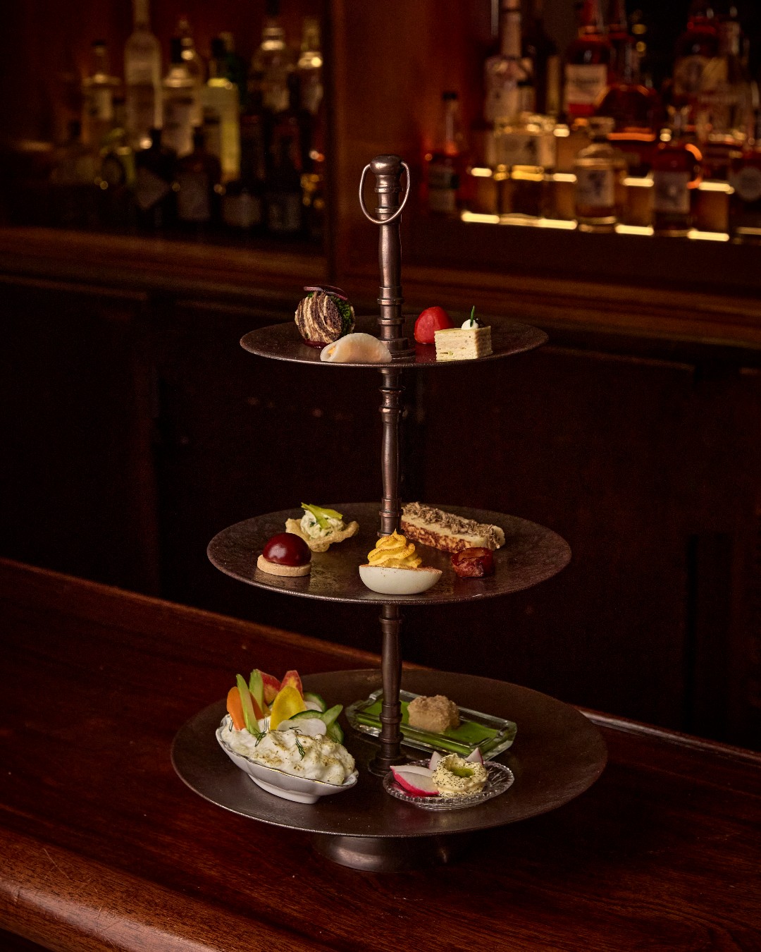 A three-tier silver stand of relish bites set on the dark wood bar at The Ives, with backlit bottles behind