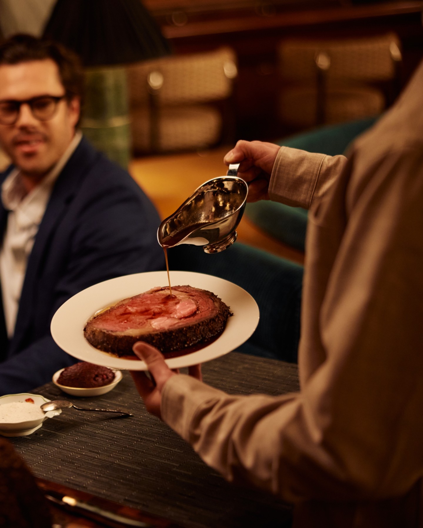 A server pours jus from a silver sauce boat over a slice of wagyu prime rib in front of a diner at The Ives