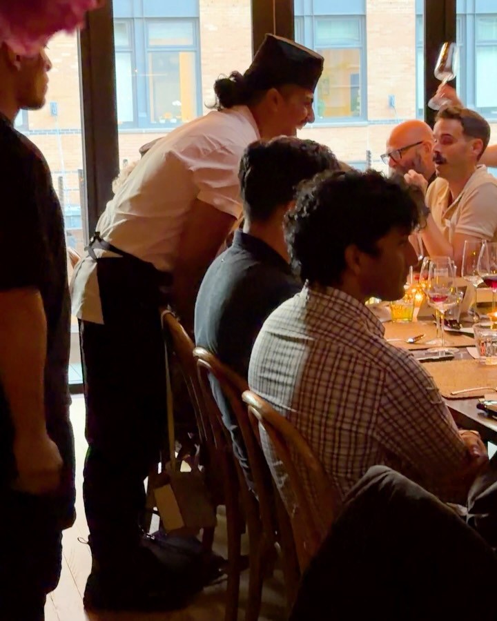Guests seated at a long communal table while a server in a white jacket delivers a course