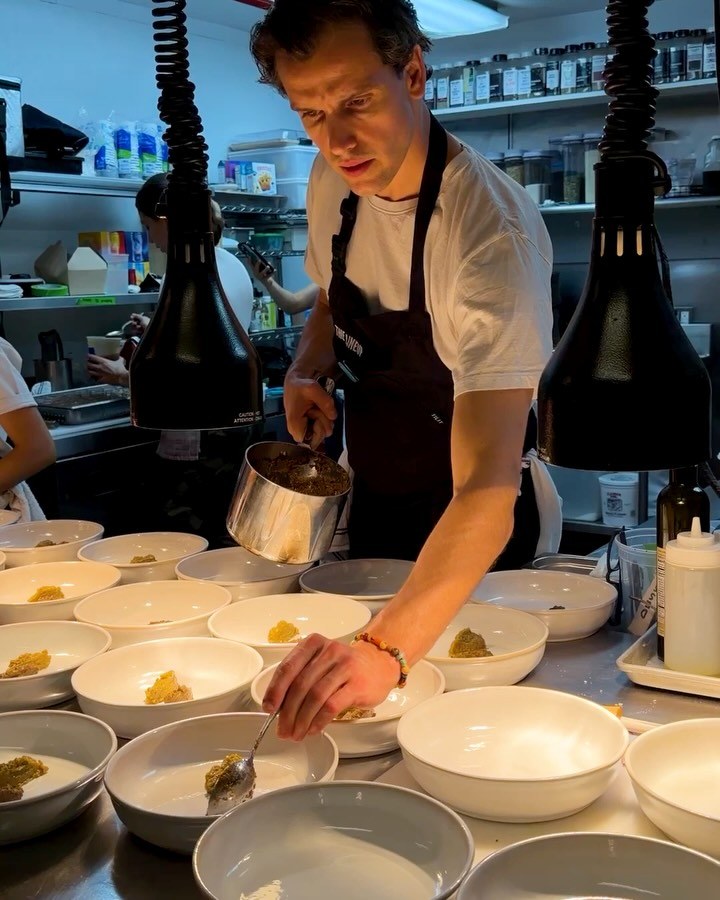 A cook in a black apron spooning sauce into a long row of white bowls at the pass