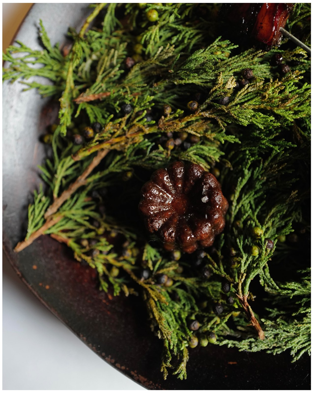 A dark lamb fat canele resting on a bed of evergreen sprigs
